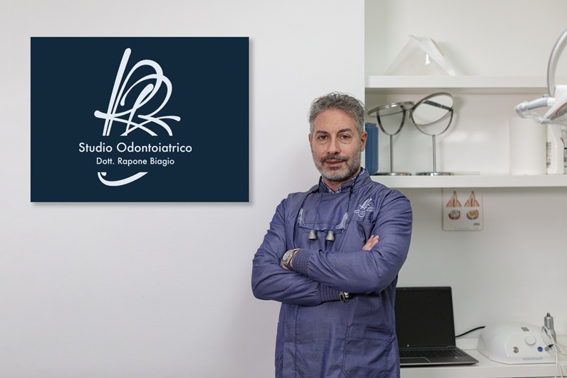 Dentista in casacca blu a braccia conserte accanto all'insegna Studio Odontoiatrico Dott. Rapone Biagio