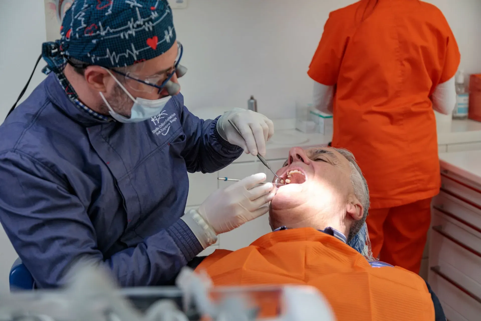 Dentista con cuffia blu e lenti ingrandenti esamina la bocca di un paziente con bavaglino arancione, Studio Rapone